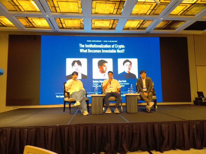 Three men sit on chairs on a stage during a panel discussion, with the screen behind them titled 'The Institutionalization of Crypto: What Becomes Investable Next?' featuring the names and portraits of panelists Robbie Huhn, Pan Park, and a third individual.
