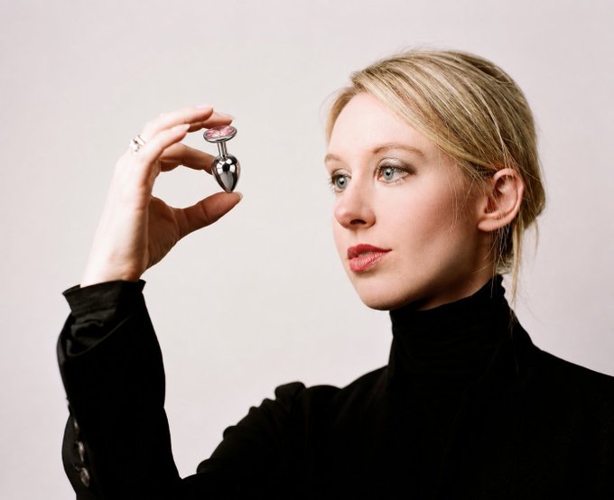 A blonde woman in a black turtleneck holds up a small heart-shaped perfume bottle, gazing thoughtfully to the side.