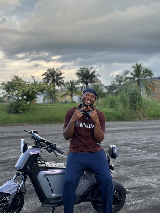 A smiling man wearing a 'HUSTLEDAILY' t-shirt and blue durag poses with both hands making gesture signs while leaning against a modern gray motorcycle in an open area with palm trees in the background.