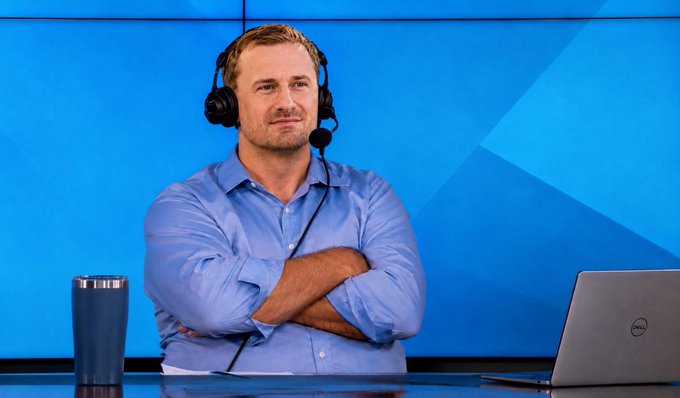 A man in a blue button-down shirt wearing a headset with a microphone, sitting at a desk with crossed arms in front of a blue backdrop, with a Dell laptop and travel mug nearby.