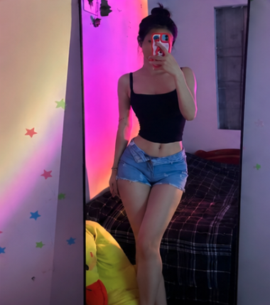 A mirror selfie of a person wearing a black crop top and denim shorts in a room illuminated with pink and purple neon lighting and star decals on the wall.