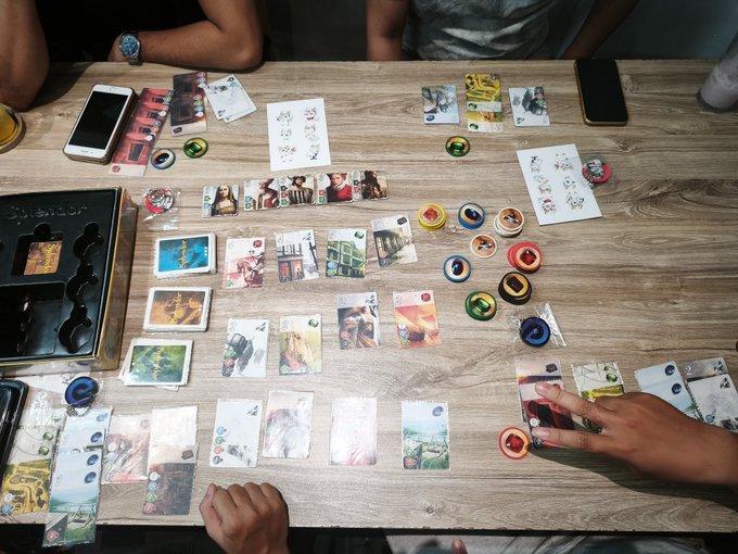 A group of people are playing a board card game on a wooden table, with various illustrated cards, colored tokens, and smartphones scattered around the play area.