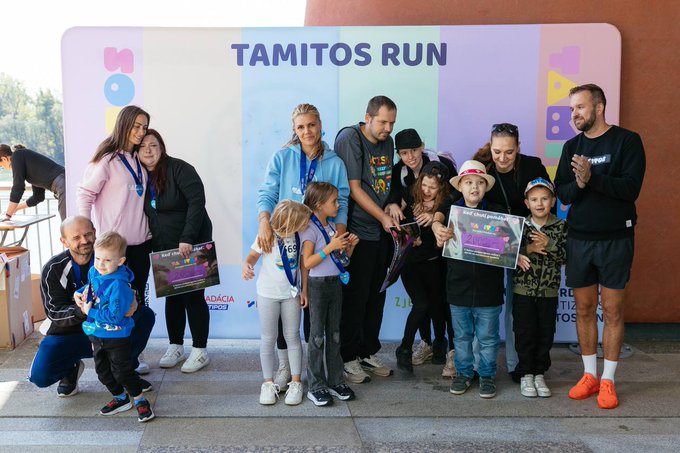 Group photo from a 'TAMITOS RUN' charity event showing adults and children posing together, with some holding certificates and a child wearing a medal, against a colorful branded backdrop.