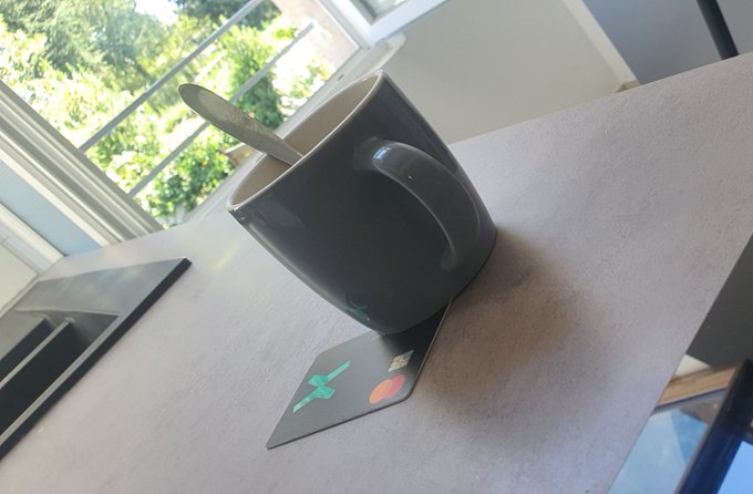 A black ceramic coffee mug with a spoon inside sits on a light-colored table next to a coaster with stickers, with a window showing greenery in the background.