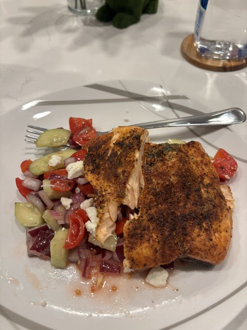 A plated meal features blackened seasoned salmon served over a fresh salad of cucumbers, tomatoes, onions, and feta cheese.