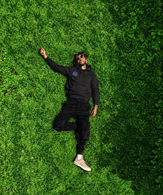 A man dressed in black with a purple chest logo lies on lush green grass, echoing the post’s “Touch grass. Clear the mind. Get back to work” message.
