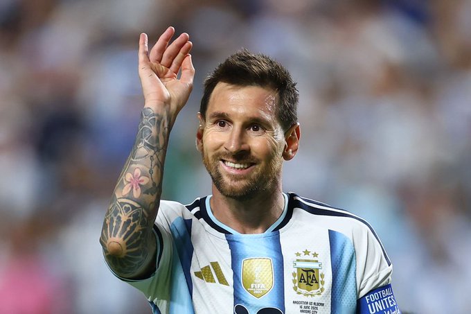 A photograph of soccer player Lionel Messi wearing an Argentina national team jersey, waving his tattooed arm and smiling during a match.