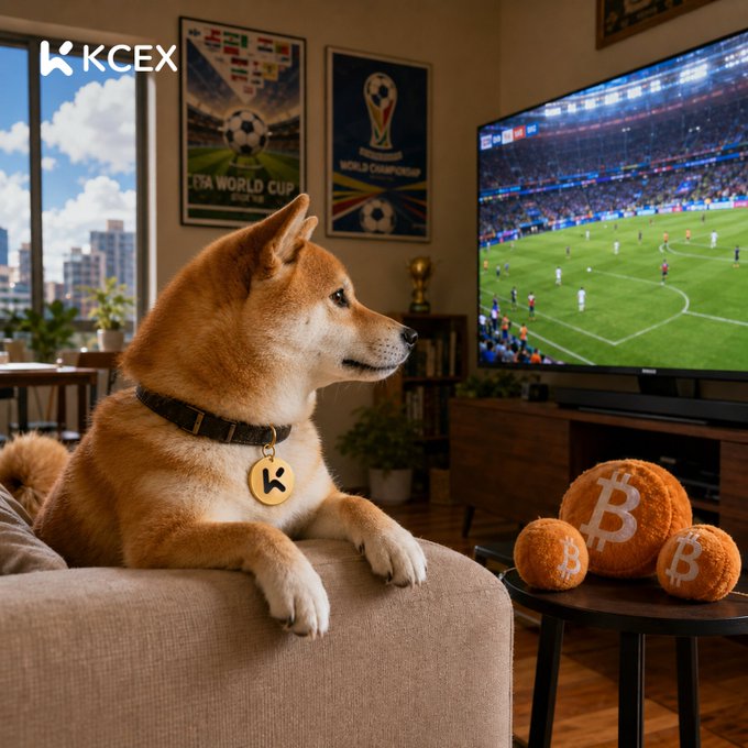 A Shiba Inu dog wearing a KCEX tag sits on a couch watching a soccer game on a large TV. Beside the dog is a small table holding three fuzzy orange balls marked with the Bitcoin 'B' symbol, and FIFA World Cup posters decorate the wall.
