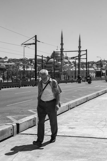A black-and-white street photograph of a man in a short-sleeved shirt walking on a sidewalk, with the Süleymaniye Mosque in Istanbul and a tram overpass visible in the background.