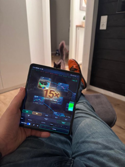 A person holding a foldable smartphone displaying a trading interface with multiple charts and a '15x' leverage indicator, with the device resting on their lap in jeans.