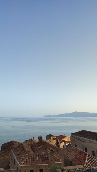 A scenic coastal view featuring terracotta-roofed buildings in the foreground overlooking a calm sea with distant mountains visible on the horizon under a clear blue sky.
