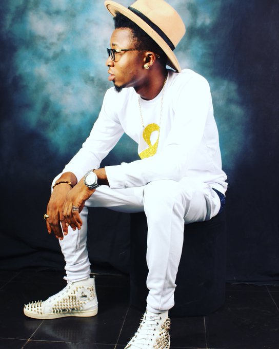 A man wearing a white outfit, a yellow ribbon shirt graphic, a tan fedora, sunglasses, and studded white sneakers sits on a black stool against a smoky blue backdrop.