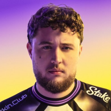 A portrait of a bearded man with curly hair wearing a black jersey with "Stake" and "Skin.Club" sponsor logos against a purple and orange gradient background.