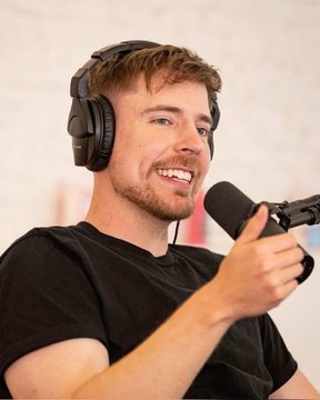 A young man with brown hair wears headphones and speaks into a microphone, smiling as he records a podcast.