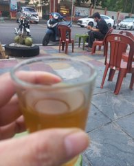 A hand holds a small glass of golden beer or tea in the foreground at an outdoor café, with patrons seated at red plastic tables and chairs on a tiled street plaza.