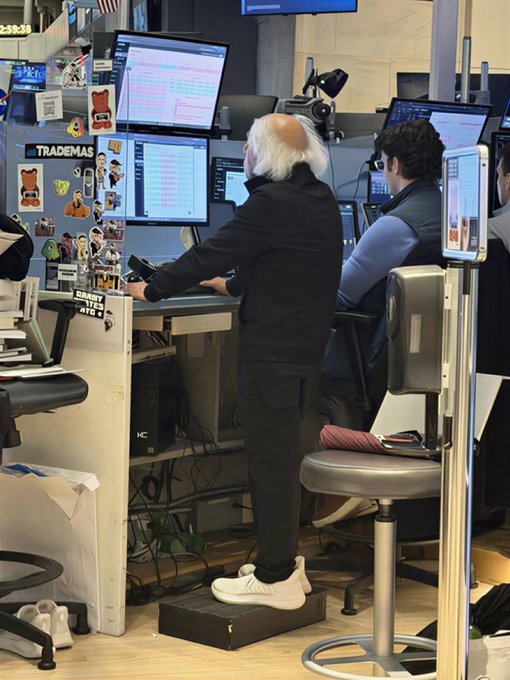 A person stands on a step stool working at a trading desk labeled 'TRADEMAS', surrounded by multiple monitors displaying financial data.
