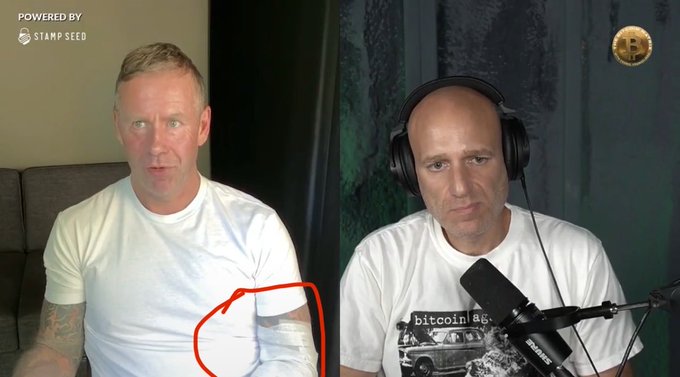 A split-screen video still featuring two men, with a 'Powered by Stamp Seed' logo in the top left and a Bitcoin logo in the top right; the man on the left wears a white t-shirt and has a red circle drawn around his shoulder.