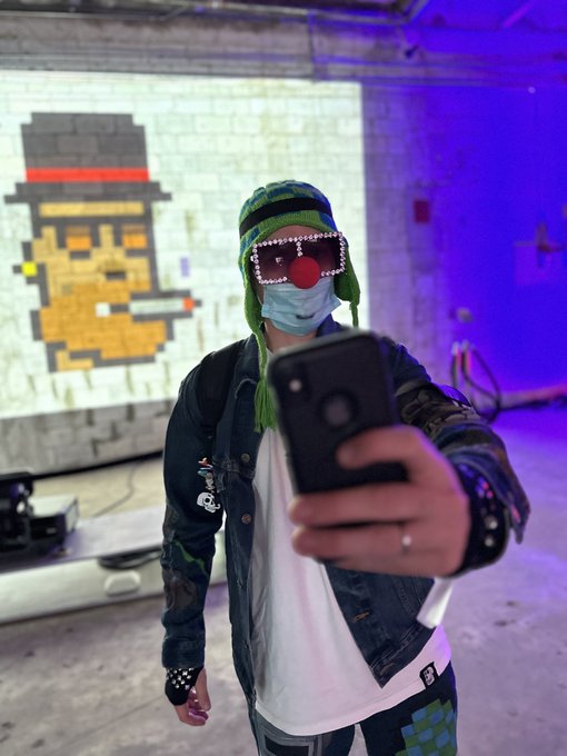 A person wearing a green knit hat, LED glasses, and a red clown nose takes a selfie with their phone in front of a pixelated duck mural illuminated with purple lighting.