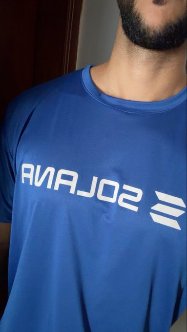 A close-up photo of a bearded person wearing a blue t-shirt with the white 'SOLANA' logo and text printed upside down (mirrored).