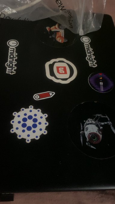 A black laptop covered with various stickers, including ones reading "...midnight" and "(?)", along with designs of a fried egg, a pencil, a virus, and a character on a bicycle.