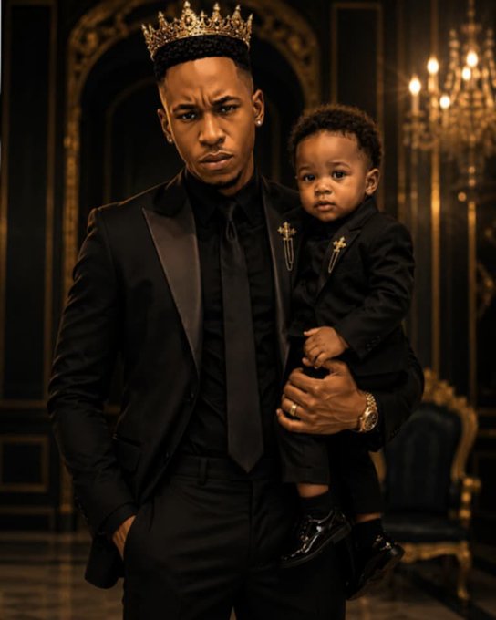 A man in a black suit and golden crown holding a young boy who is also dressed in black formal attire with cross pins, posed in an ornate room with a chandelier.