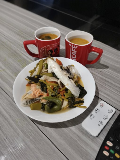 A meal of fish and vegetables in a white bowl sits on a wooden table next to two red mugs filled with hot beverages and a TV remote control.
