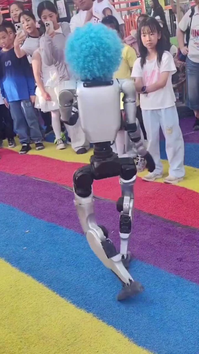 The image features a robot with blue hair, walking across a colorful carpeted floor. A crowd of people surrounds the robot, some taking pictures while others look on with interest. The caption reads "Me when I find the person who told me to buy bitcoin at $120k".