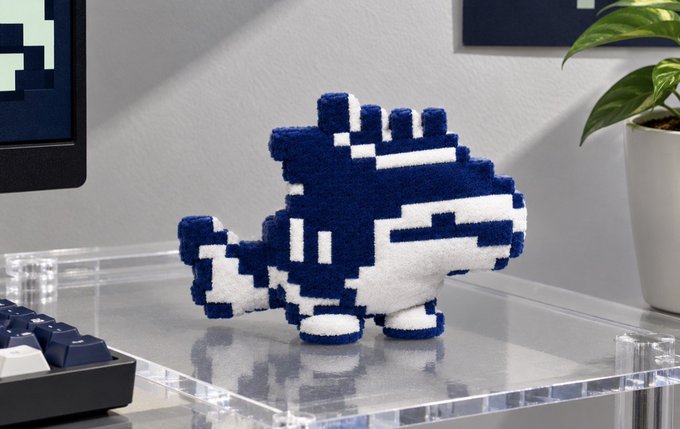 A blue and white pixel-art style figure resembling a small character with a crown sits on a clear acrylic stand next to a keyboard and plant.