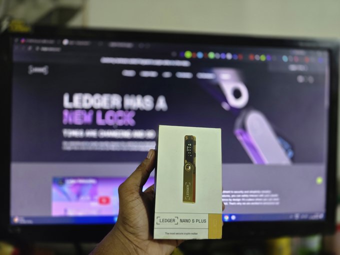 A hand holds up the packaging for a Ledger Nano S Plus hardware wallet in front of a computer screen displaying "LEDGER HAS A NEW LOOK" on the Ledger website.