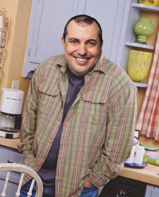 A man with short dark hair, smiling broadly, wears a green and red plaid flannel shirt over a purple t-shirt in a cozy kitchen setting with a coffee maker and decorative vases. The relaxed, casual home environment and warm smile contrast humorously with the post's irreverent caption about "setting the pleb slop in motion."