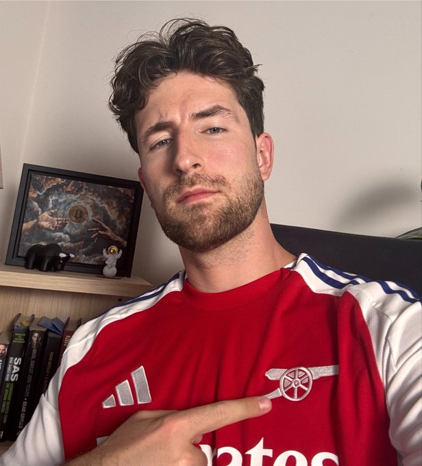 A man with dark curly hair and a beard is taking a selfie while wearing a red and white Arsenal football jersey with the Adidas logo and cannon crest visible, pointing at the team badge with his finger.