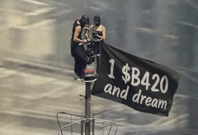 Two figures in balaclavas atop a pole holding a black flag that reads "1 $B420 and dream".