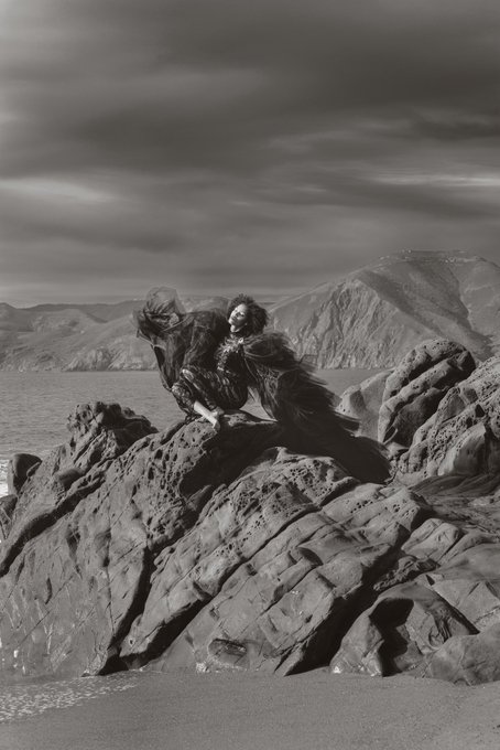 A black-and-white photograph shows a figure in flowing dark clothing reclining on a large rocky outcrop by the sea, with mountains and a dramatic cloudy sky in the background.