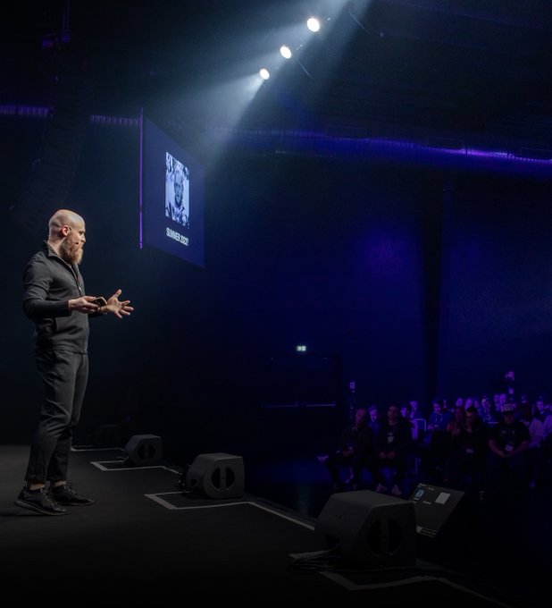 A bearded presenter in black attire speaks on a dimly lit stage to an audience, with a large screen behind him displaying "SCANDER BEG" beneath a portrait. The image captures a live trading seminar setting where the speaker is likely delivering the message about distinguishing between emotional risk tolerance and financial risk capacity.