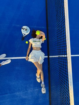 An aerial view of a padel or tennis player in a white outfit on a blue court, holding a racket while a yellow ball is in mid-air near her.
