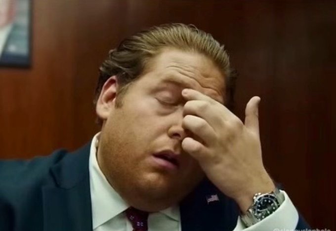 A man in a suit with an American flag pin rubs his forehead with his eyes closed, wearing a watch, appearing weary or exasperated.