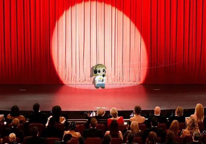 A cartoon baby character wearing a white t-shirt stands center stage under a spotlight, in front of red curtains, addressing an audience seated in a theater.