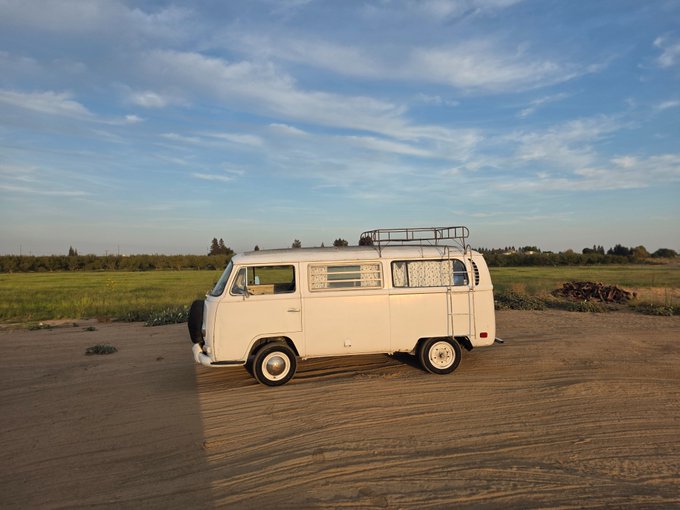 A classic white Volkswagen camper van parked on a dirt area at golden hour, with green fields and a cloudy blue sky in the background.