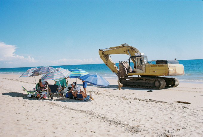 A surreal beach scene where people lounge under colorful umbrellas on the sand while a large yellow excavator operates right next to them by the ocean.