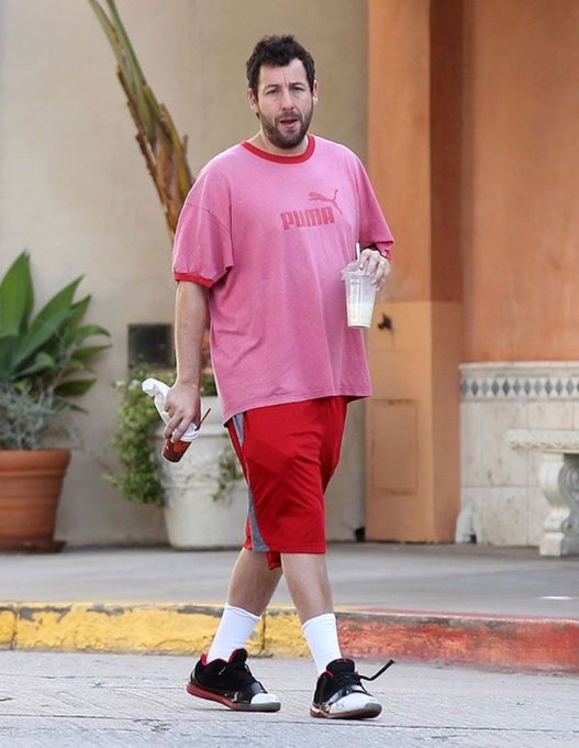Adam Sandler walks outdoors wearing a pink PUMA t-shirt, red shorts, white socks, and black and white sneakers, carrying a water bottle and a drink cup.