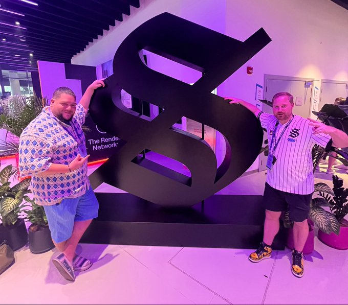 Two men pose playfully on either side of a large black sculpture with "The Render Network" text at the bottom, in a modern indoor space with purple lighting.