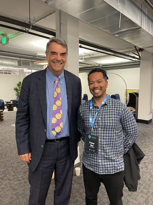 Two men stand smiling at what appears to be a conference; the man on the left wears a suit with a distinctive purple tie patterned with Bitcoin logos, while the man on the right wears a plaid shirt with a 'Parknr Hub' lanyard.