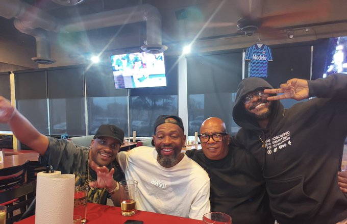 Four men posing together at a restaurant/sports bar with TVs and a framed Modelo jersey in the background.
