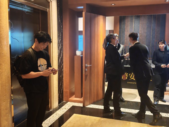 Several people in business suits and one in a skull t-shirt gather in a marble-floored lobby near a door labeled 'CHANGPE'.