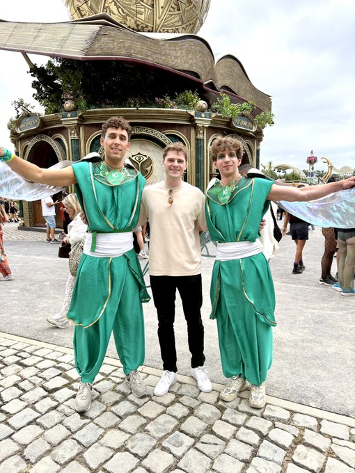 A young man poses between two performers dressed in green genie-like costumes at Tomorrowland, with a sign reading "TOMORROWLAND" visible on the ornate building behind them. The scene captures a fun theme park moment sponsored by KuCoin, matching the post's shoutout to the crypto exchange.