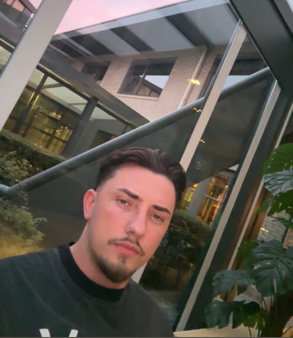 A selfie of a young man with dark hair and a goatee, wearing a black shirt, taken indoors near large windows.