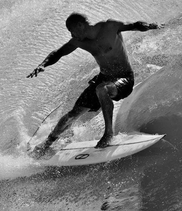 A black-and-white photograph of a muscular man surfing on a wave, balancing on his board with arms outstretched.