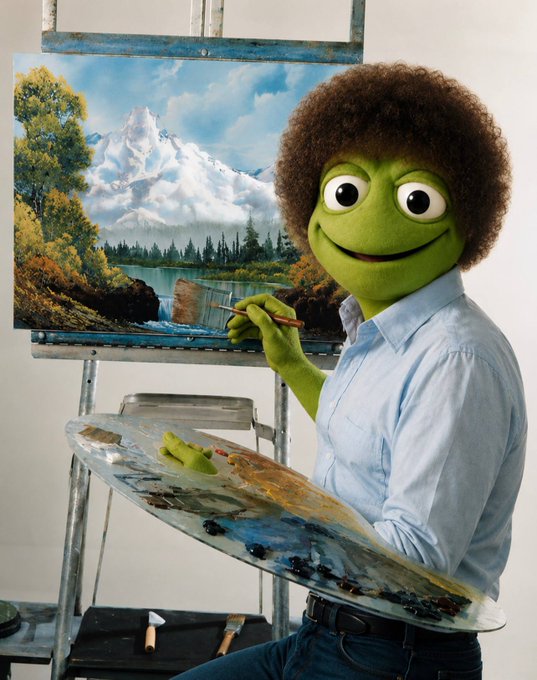 A green Muppet-like frog character with an afro and painter's palette stands at an easel, adding finishing touches to a serene mountain landscape painting. The image visually ties to the Art Contest announcement by symbolizing the celebration of artistic creativity, with the painterly scene serving as a whimsical representation of the winning artwork and talented artists recognized in the contest.