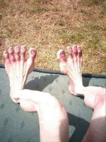 A first-person view of a person's bare feet with toes painted pink resting on a cushion outdoors on the grass.