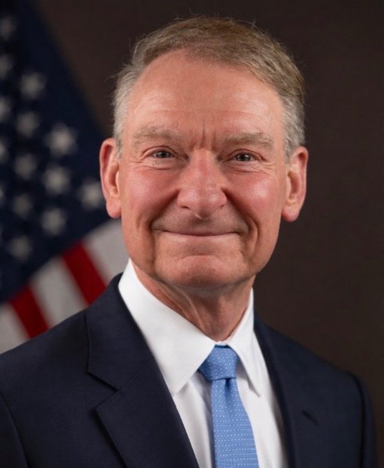 An official portrait of an older man with gray hair, smiling slightly, wearing a dark suit, white shirt, and blue patterned tie, with an American flag visible in the background.